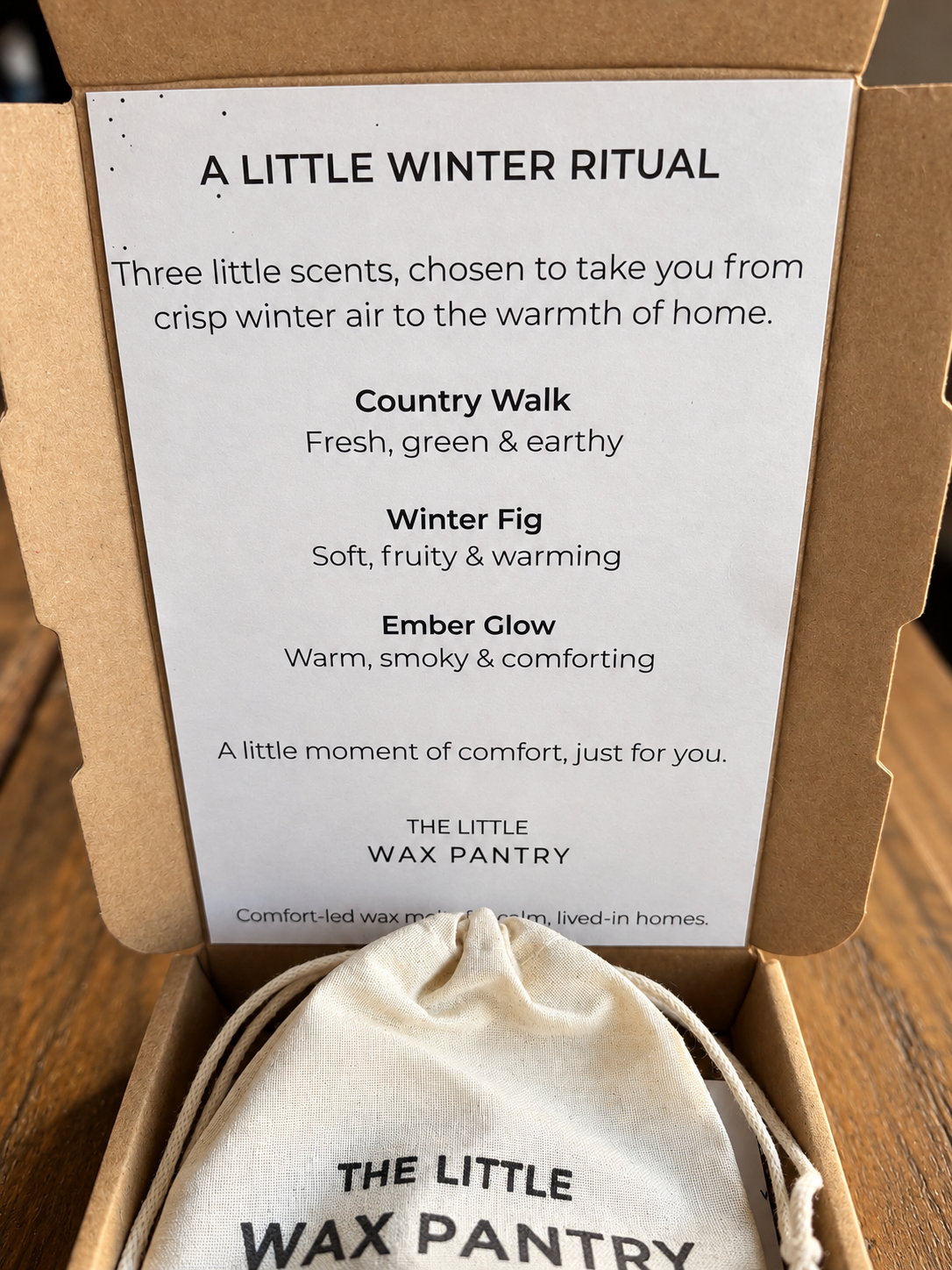 An open kraft gift box containing a white printed card titled 'A LITTLE WINTER RITUAL' with scent descriptions, and a small cream drawstring pouch printed with 'THE LITTLE WAX PANTRY' at the bottom.
