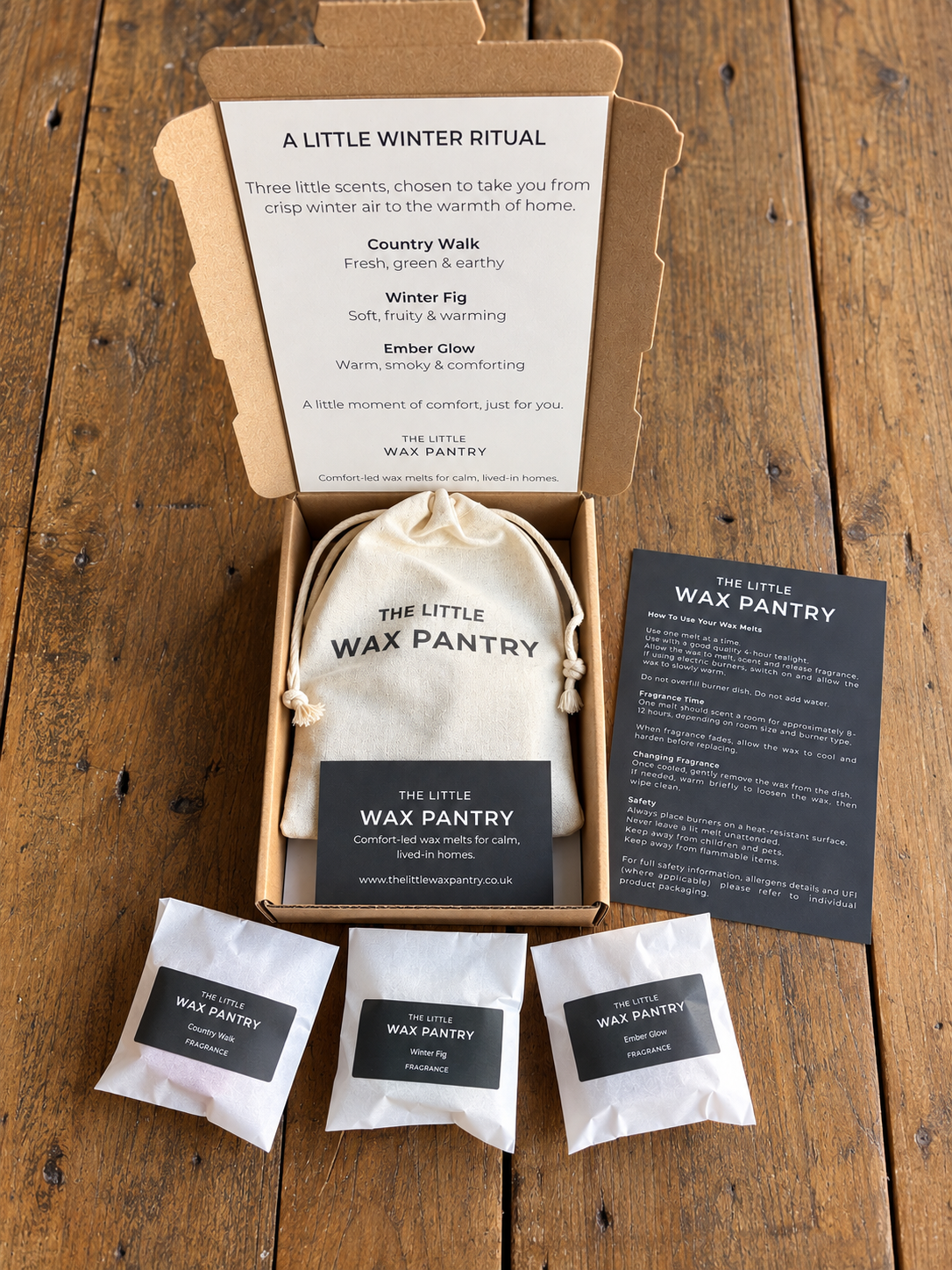 A curated gift set from The Little Wax Pantry arranged on a rustic wooden table. The set includes an open kraft gift box with a printed card, a small cream drawstring bag, three small white sachets with black labels, and a larger black printed card.
