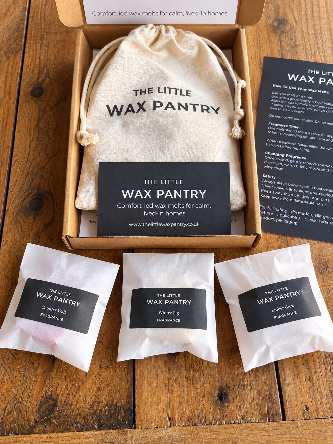 The Little Wax Pantry gift set arranged on a wooden table, featuring a kraft box with a cream drawstring bag, a black brand card, a printed instruction sheet, and three small wax melt packets labeled 'The Little Wax Pantry'.