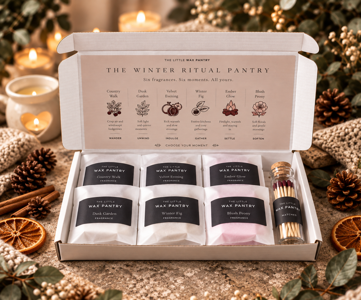 An open white gift box labeled 'The Winter Ritual Pantry' by The Little Wax Pantry, containing six small wax melts in white and pink packets with black labels and a small bottle of matches. The box is styled on a rustic surface with candles, pinecones, cinnamon sticks, dried orange slices, and greenery in the background.